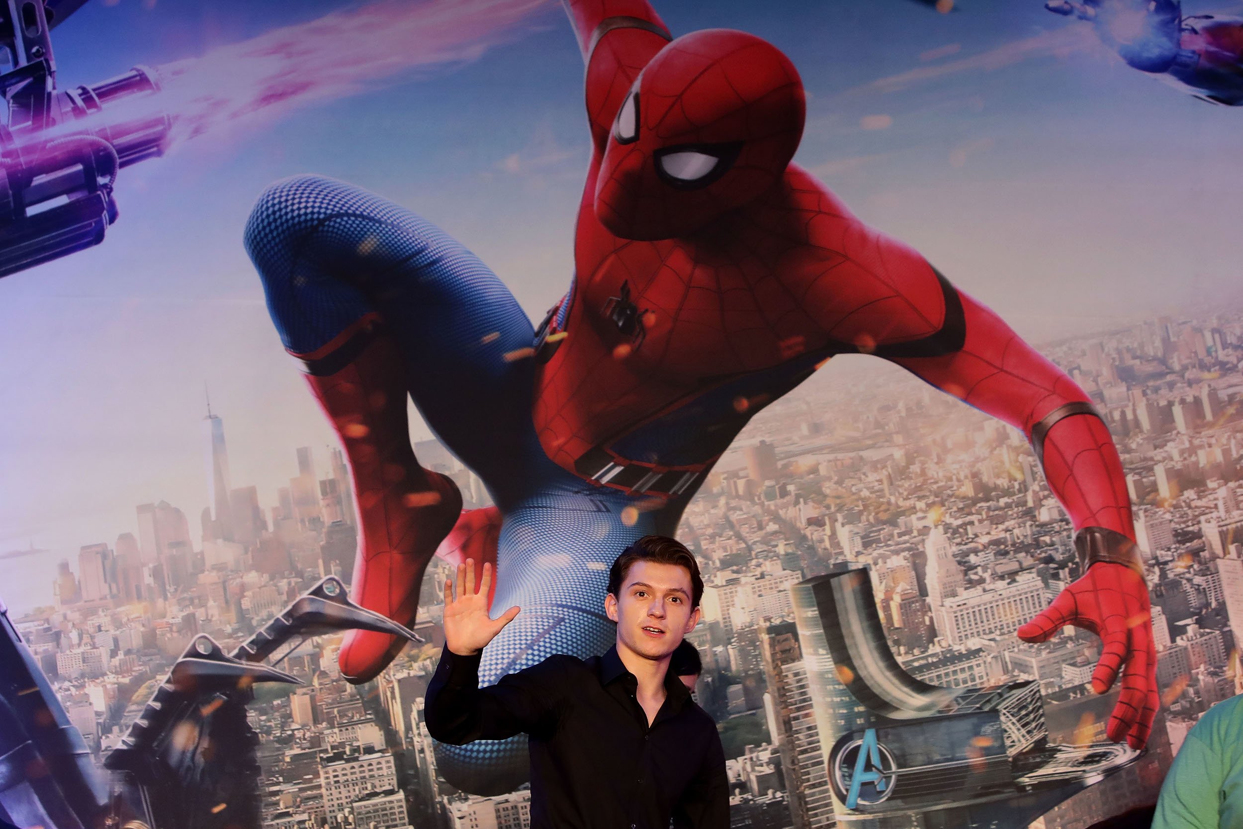 'Spider-Man: No Way Home' star Tom Holland standing beneath a Spider-Man image and waving