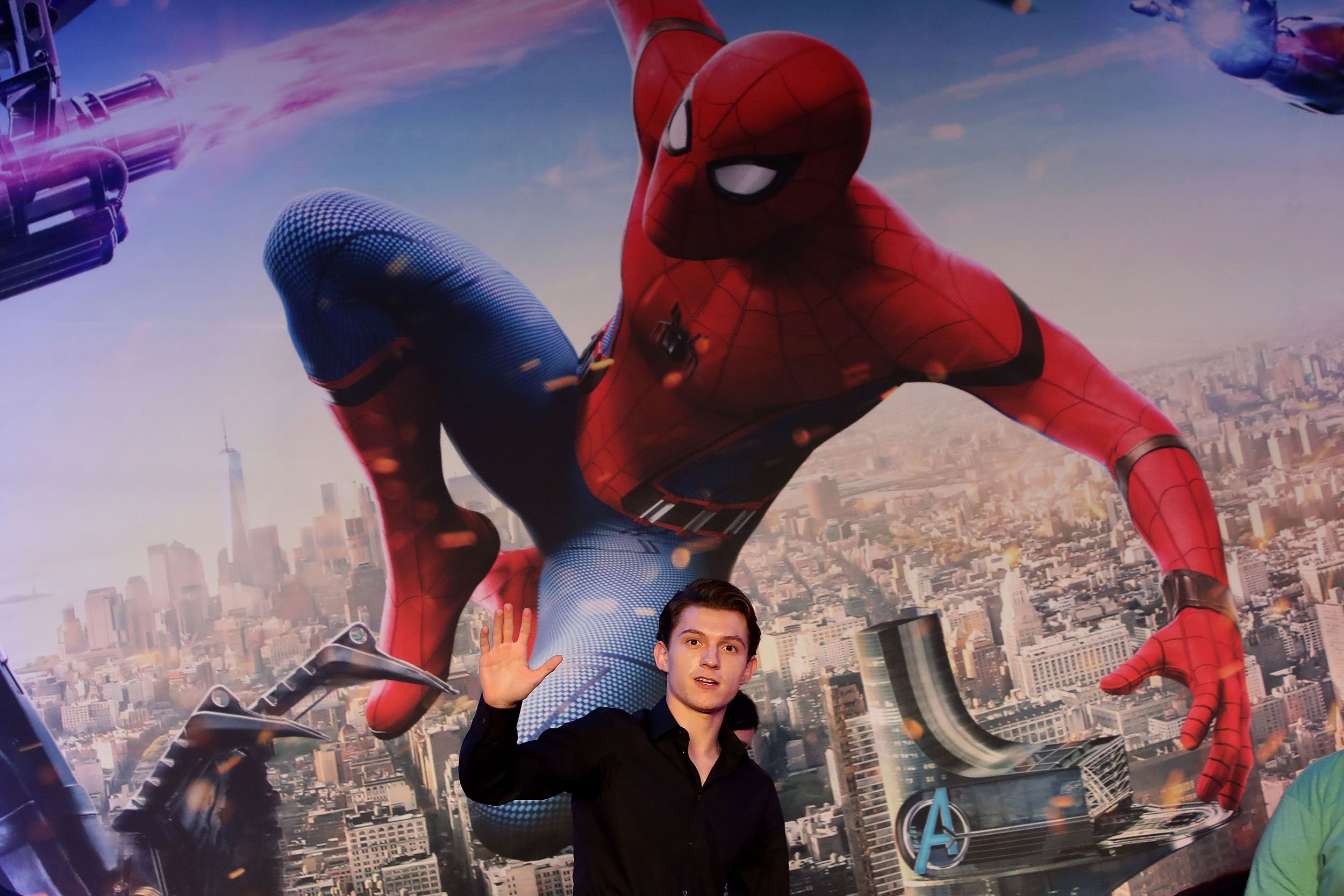 Spider-Man: No Way Home star Tom Holland standing beneath a Spider-Man image and waving