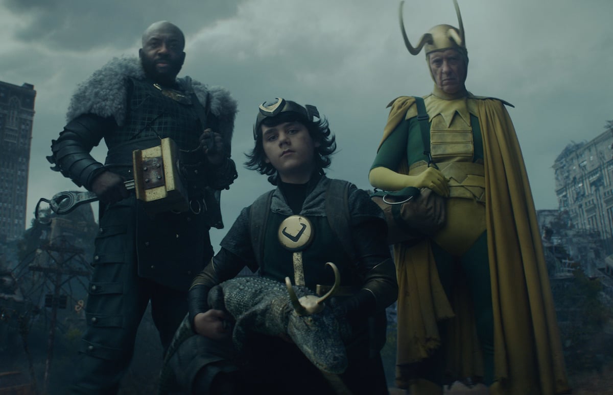(L-R) Deobia Oparei, Jack Veal, and Richard E. Grant in 'Loki'. The actors are dressed in various green and gold and black Loki costumes. The center Loki holds an alligator Loki in his arms. They stand in front of a post-apocalyptic wasteland.