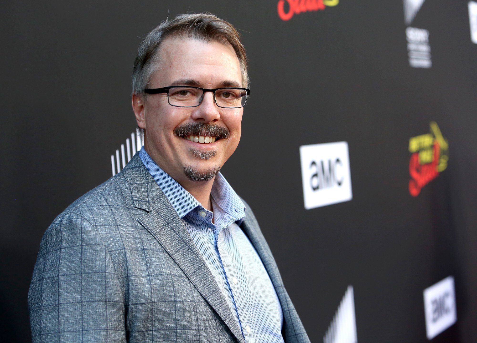 Breaking Bad and Better Call Saul creator Vince Gilligan wearing a blue button-up shirt and dark suit and standing in front of a black wall with 'AMC' written on it