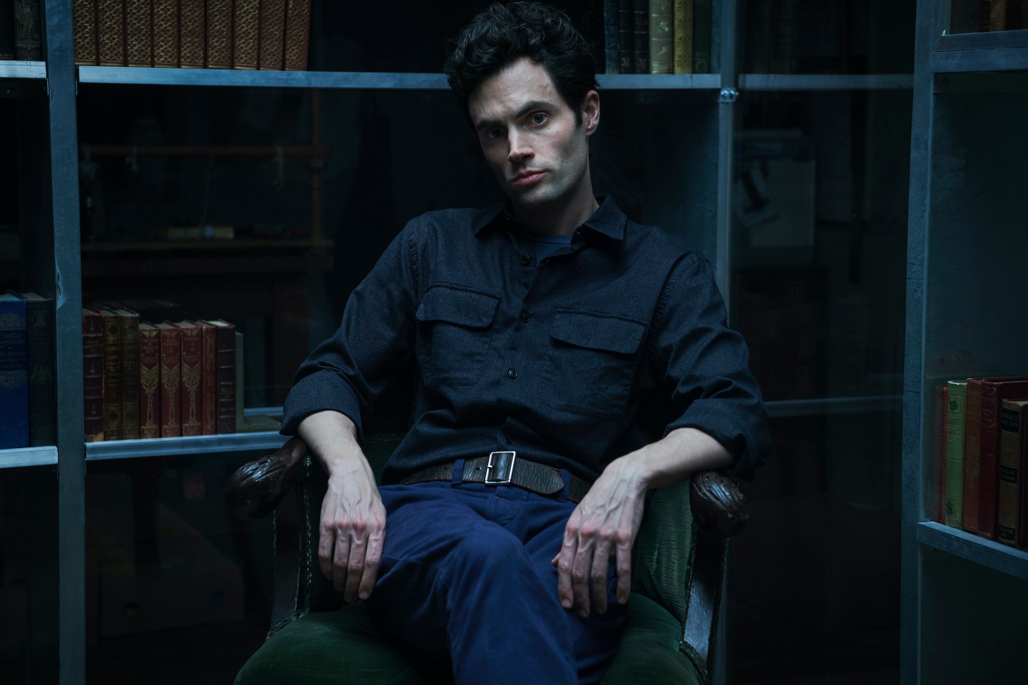 Penn Badgley as Joe Goldberg sitting in front of bookshelves