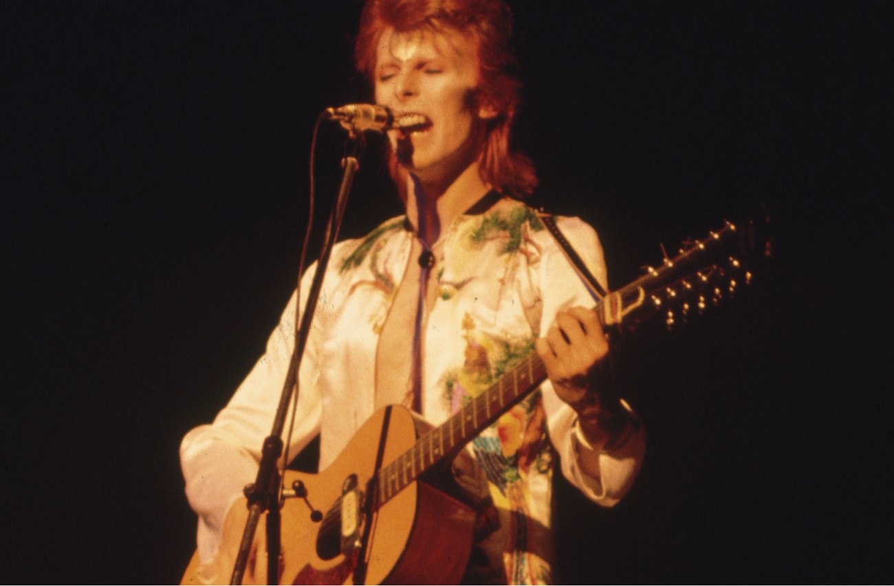 David Bowie onstage playing acoustic guitar. He wears baggy white pants, a printed shirt and has long, dyed red hair.