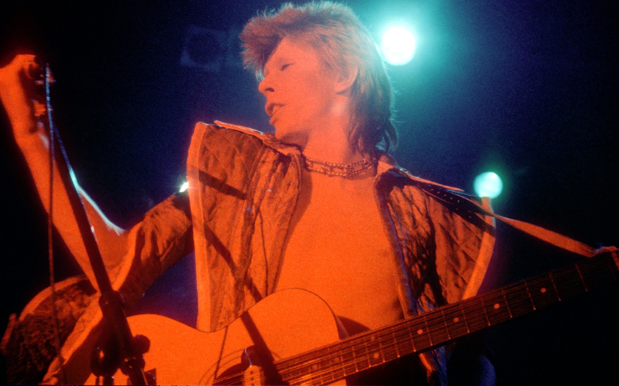 David Bowie performing under a red light with a guitar strapped across his back in 1973.