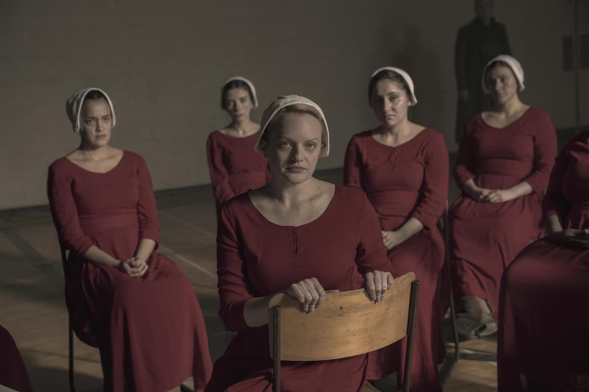 Brianna (Bahia Watson) and June (Elisabeth Moss) in 'The Handmaid's Tale' Season 3. Five women sit in school desk chairs in an old gymnasium. They wear red Handmaid dresses and white bonnets.