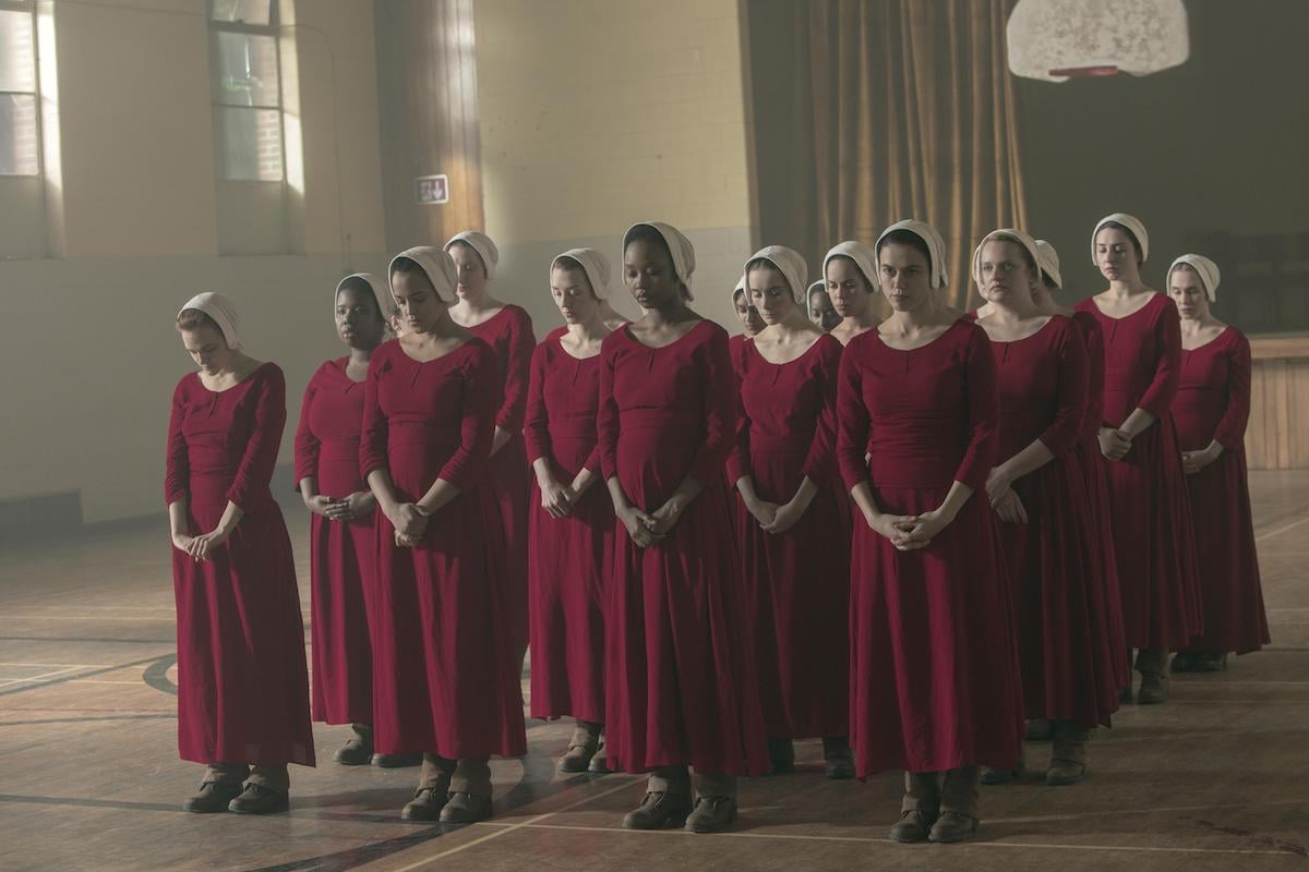 Janine (Madeline Brewer), Brianna (Bahia Watson), Ofmatthew (Ashleigh LaThrop), Alma (Nina Kiri), and June (Elisabeth Moss) in 'The Handmaid's Tale' Season 3. A group of about 20 women dressed in red Handmaid dresses and white bonnets stand in five lines with their feet together and hands crossed in front of them. They stand in an old school gymnasium.