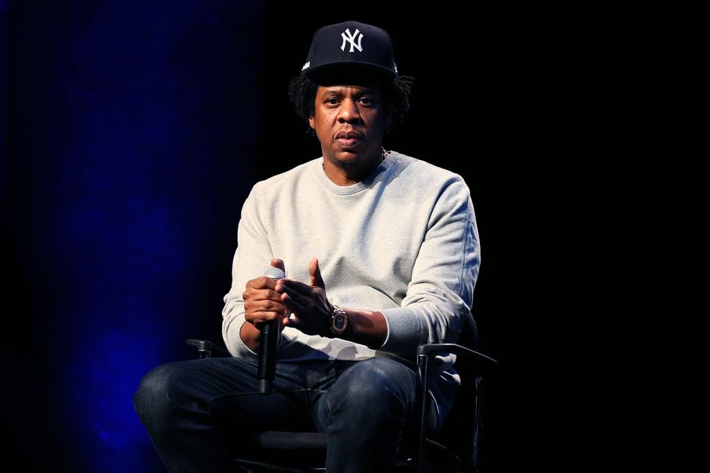Jay-Z sits on stage wearing a Yankees hat