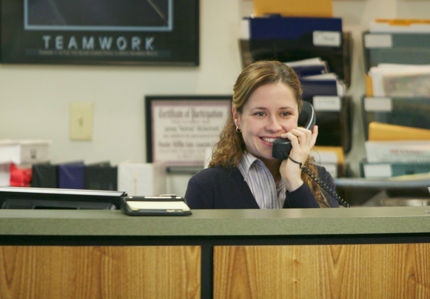 The Office star Jenna Fischer as her character Pam, smiling at the reception desk at Dunder Mifflin