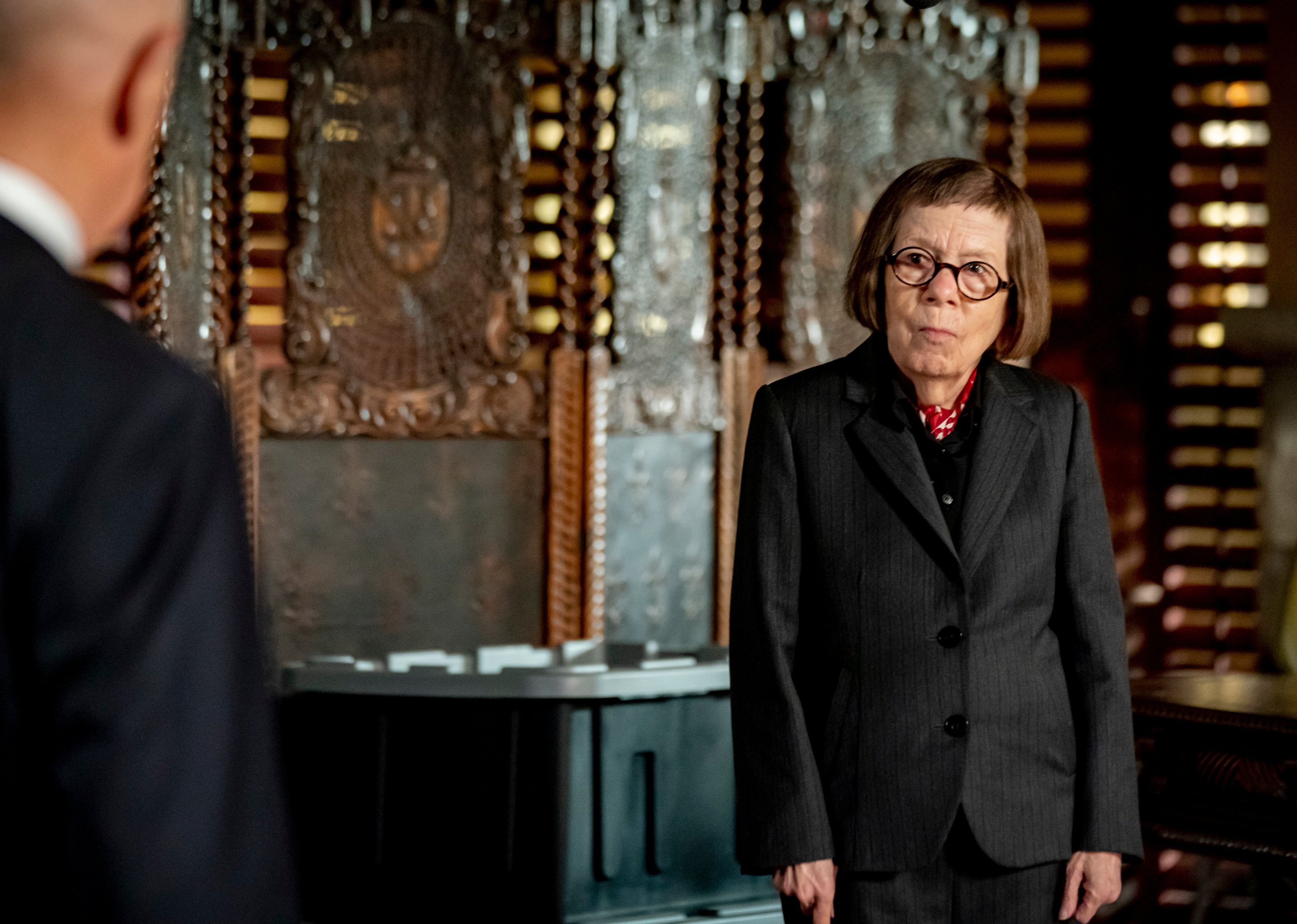 Linda Hunt wears a suit and stands in her office, looking concerned.