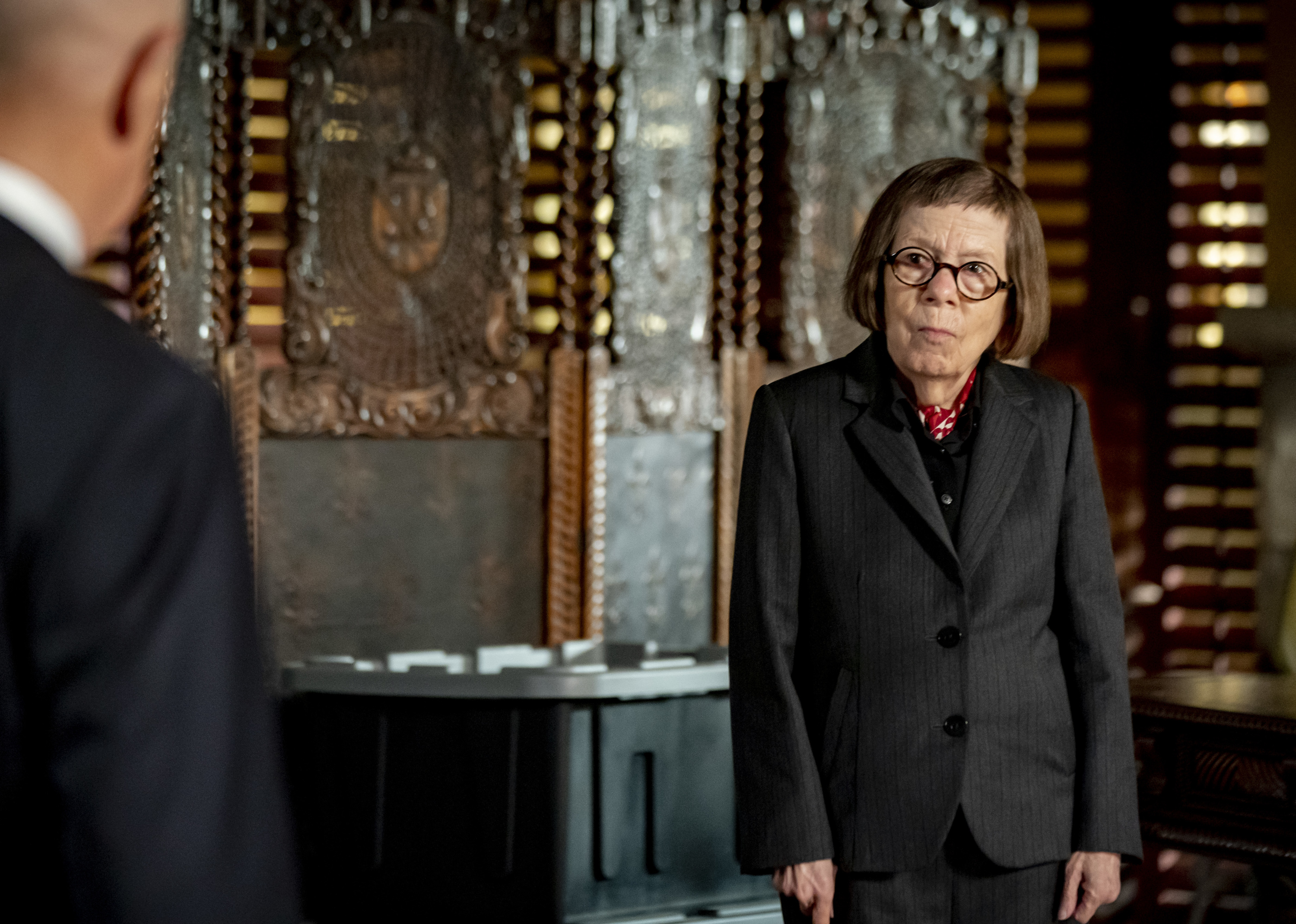 Linda Hunt wears a suit and stands in her office, looking concerned.