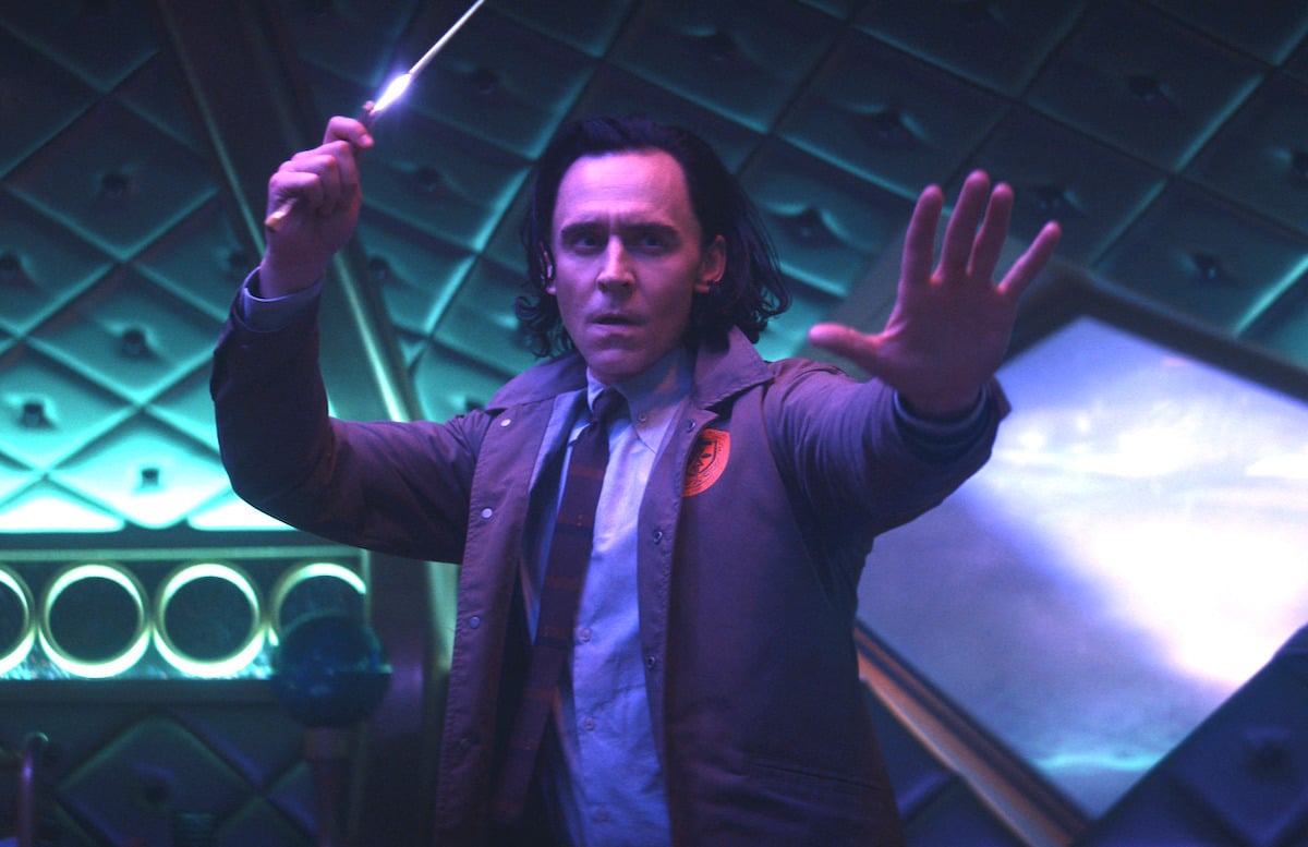 Tom Hiddleston wields a dagger while wearing a shirt and tie with a brown TVA jacket in a room lit with purple and teal lighting.