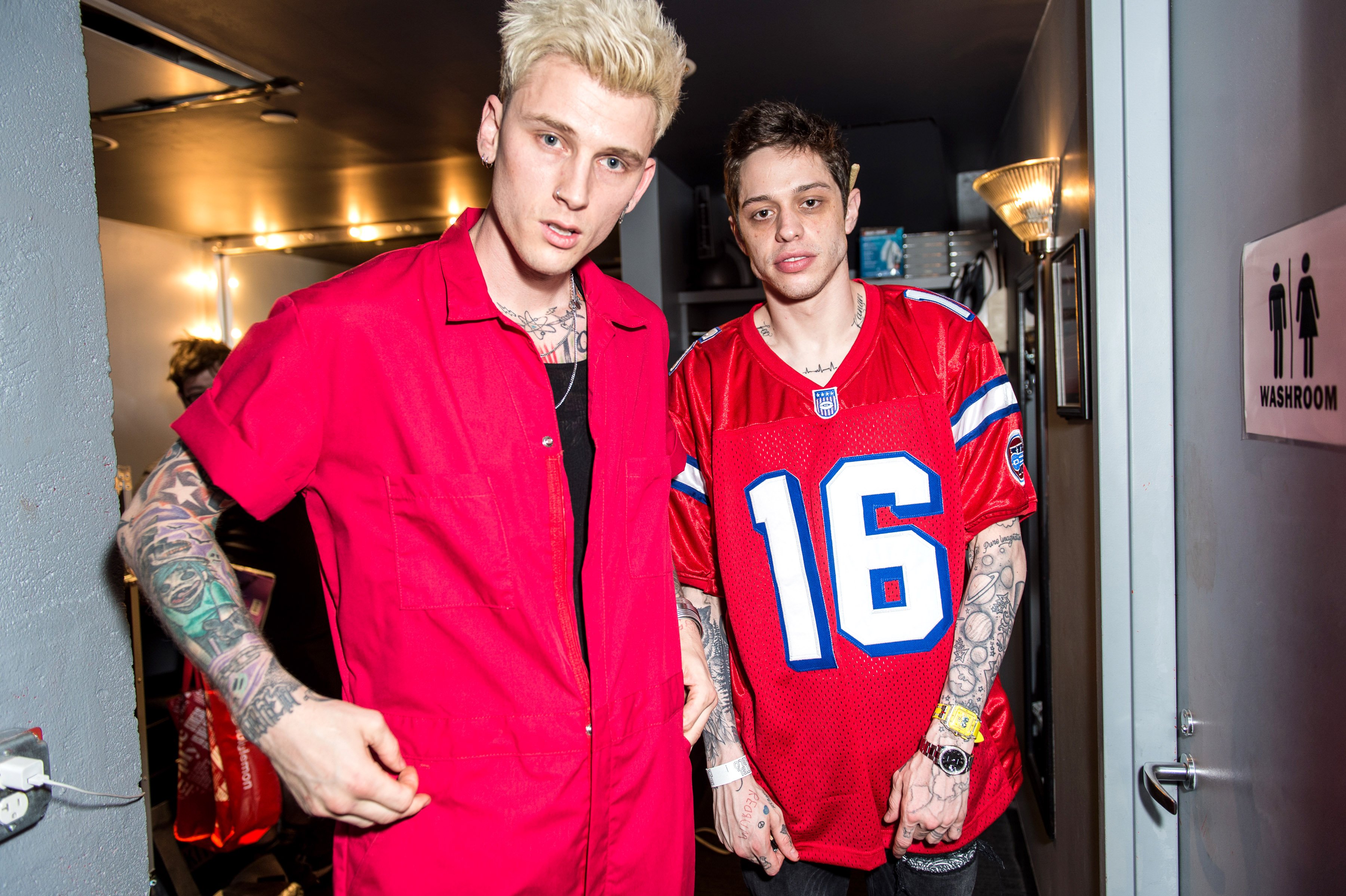 Machine Gun Kelly and Pete Davidson stand in a hallway outside a bathroom door while wearing red outfits.