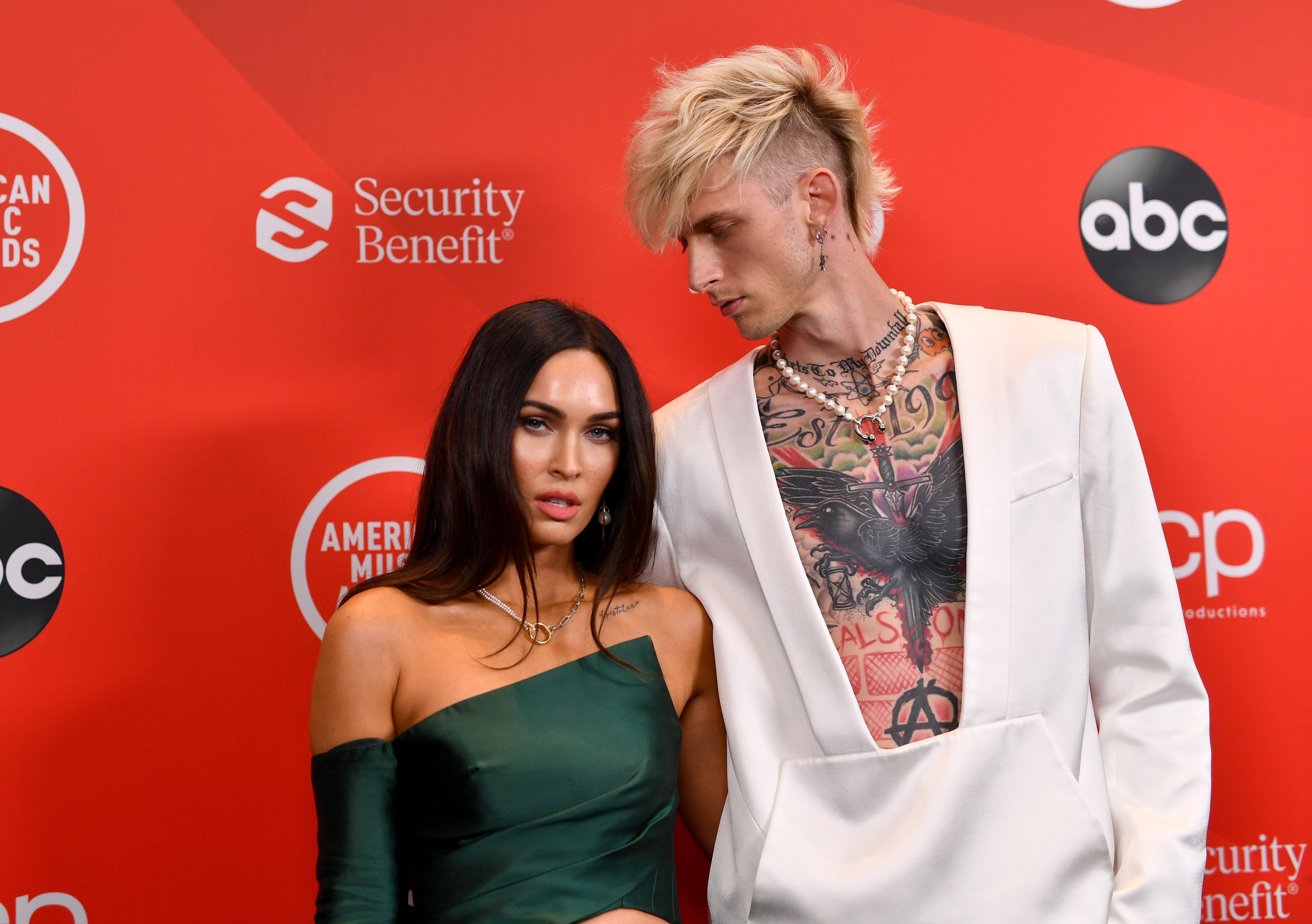 Megan Fox wears a green dress while standing with Machine Gun Kelly, who is wearing a white suit.
