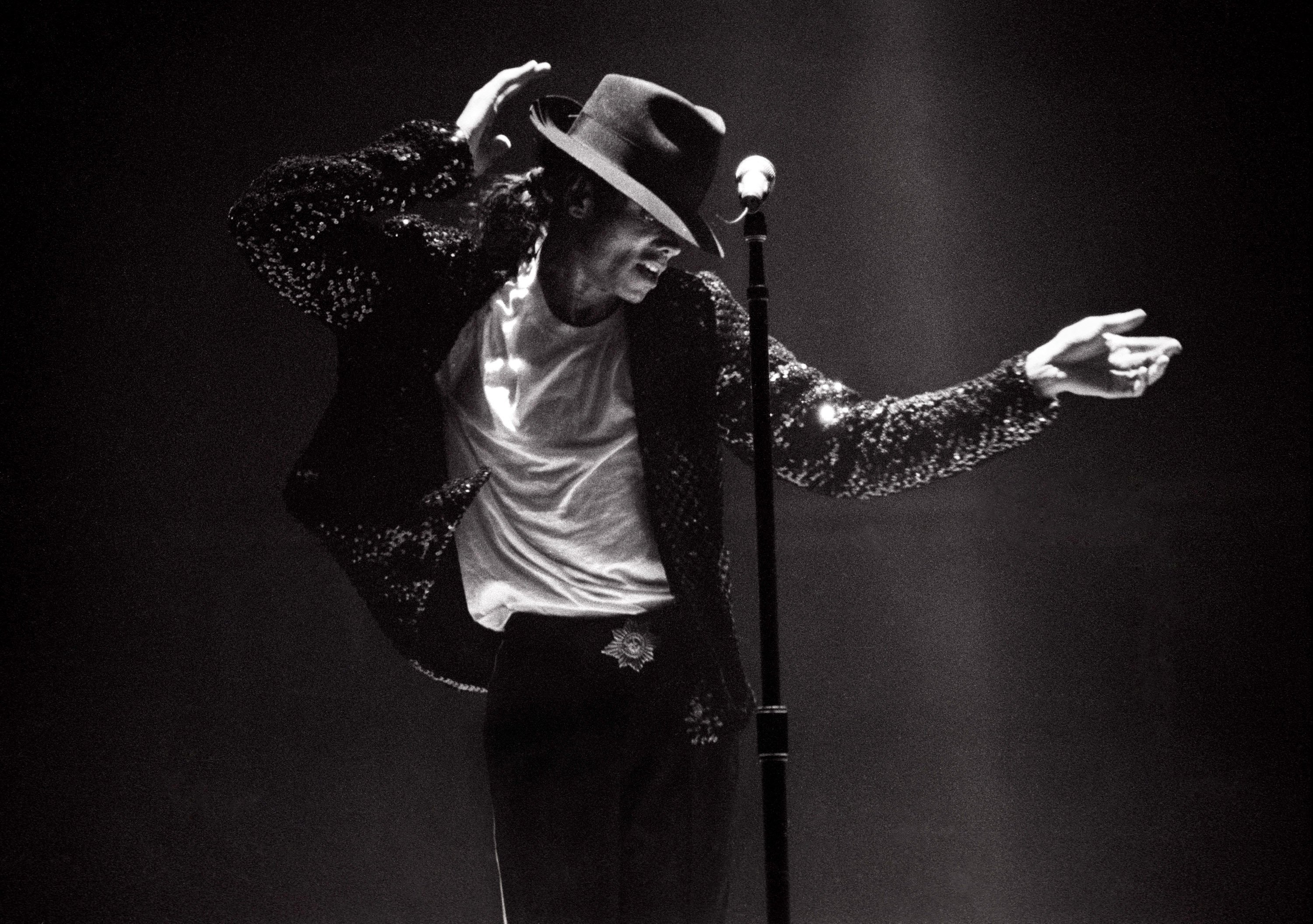 Michael Jackson wearing a fedora and a sparkly jacket while doing the moonwalk in front of a microphone