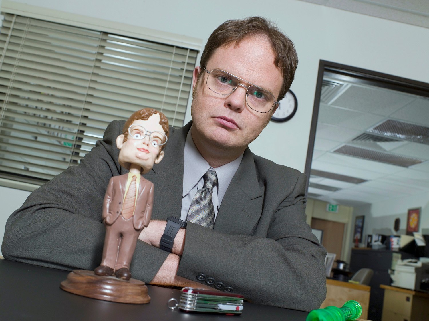 The Office star Rainn Wilson as character Dwight Schrute sitting behind a bobblehead Dwight doll.