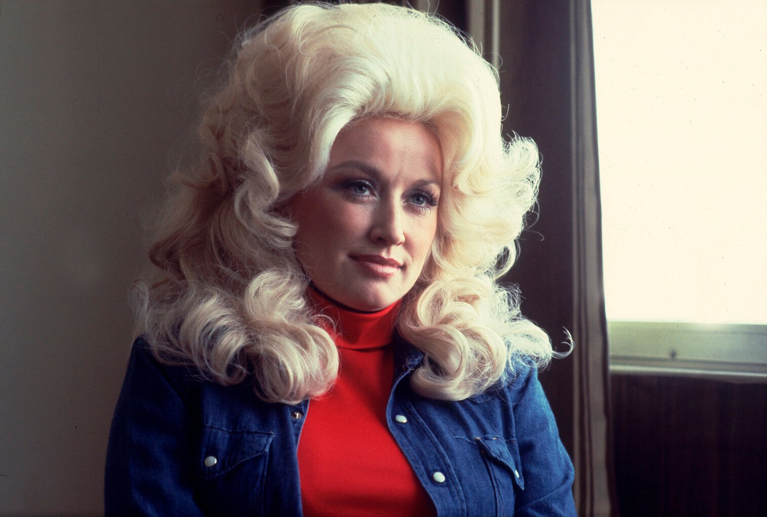 Portrait of Dolly Parton at the Holiday Inn in Chicago, Illinois, April 30, 1977. She's wearing her hair big and blonde. She has a red top on and a jean jacket.