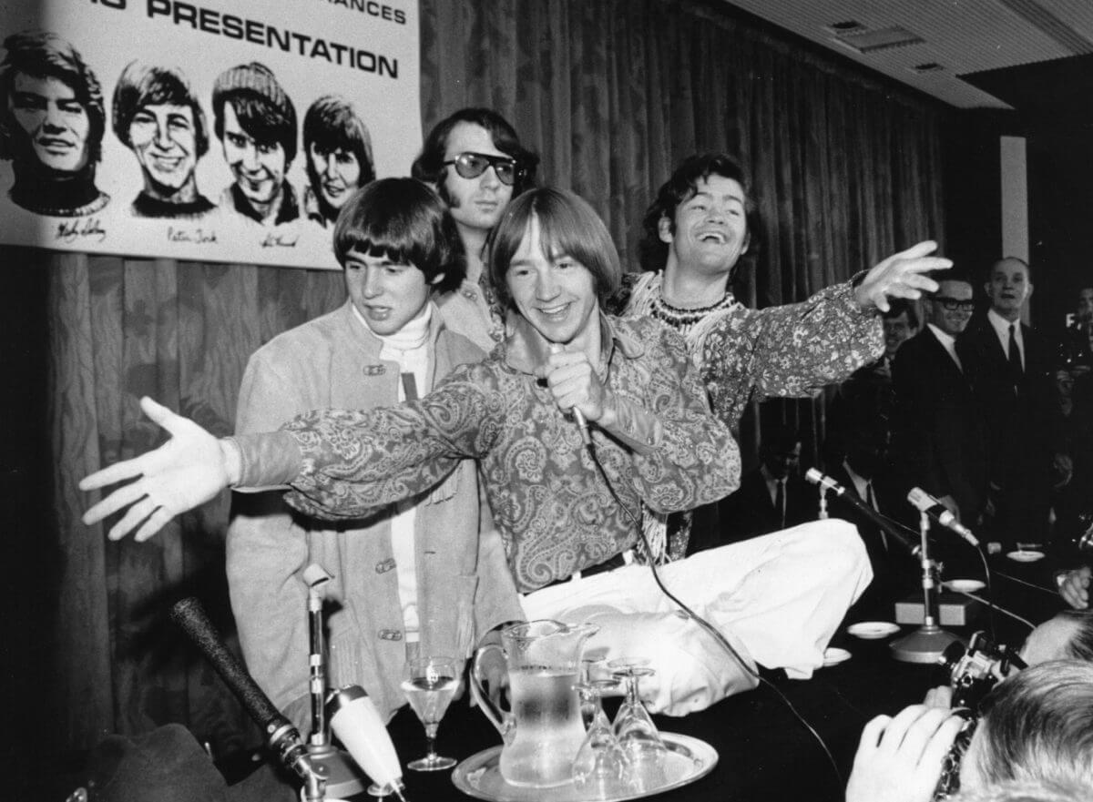 The Monkees stand behind a table with a water pitcher on it. 