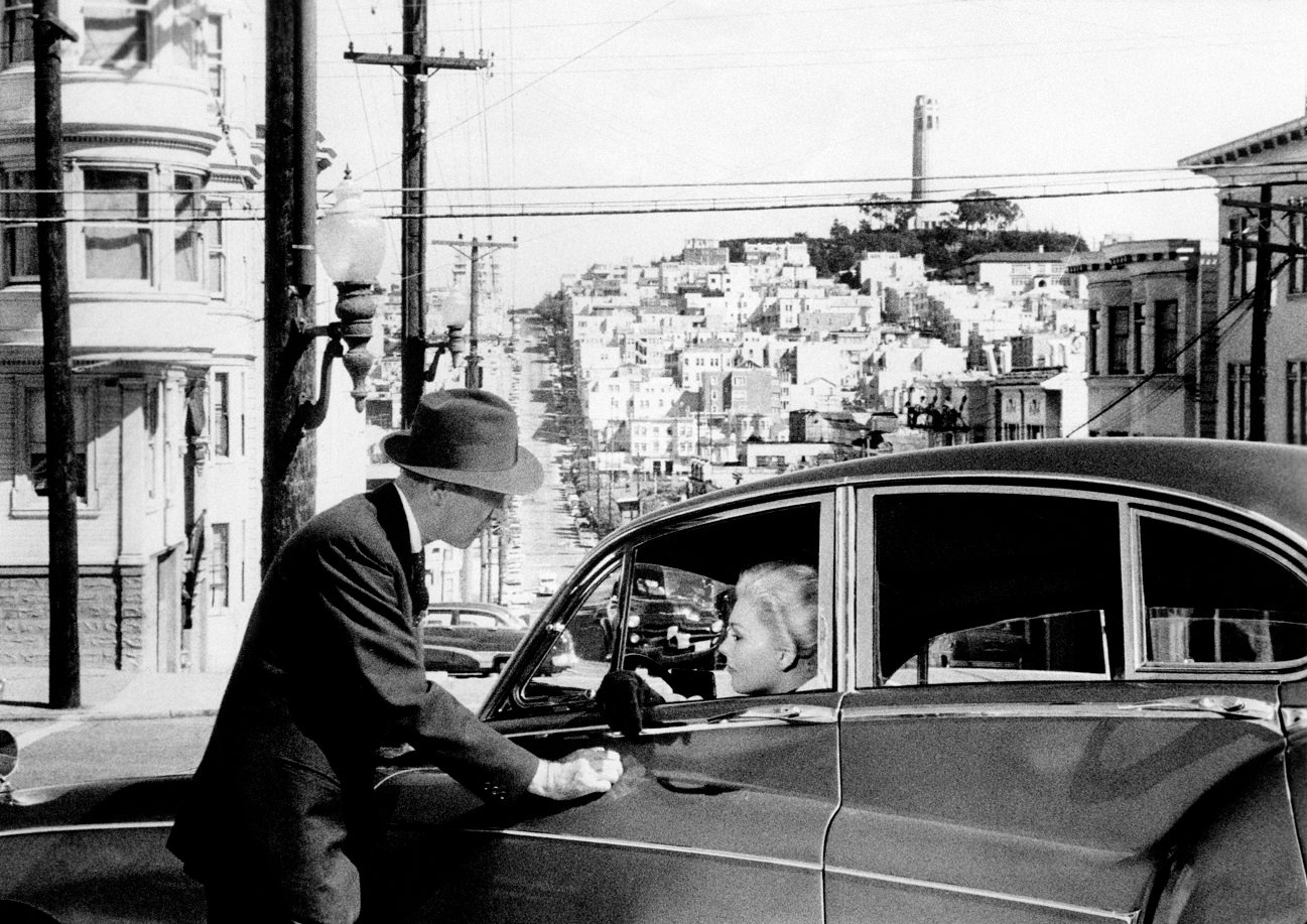 Jimmy Stewart leans on a car in which Kim Novak sits in the driver's seat with a view of San Francisco in the background. From 'Vertigo'