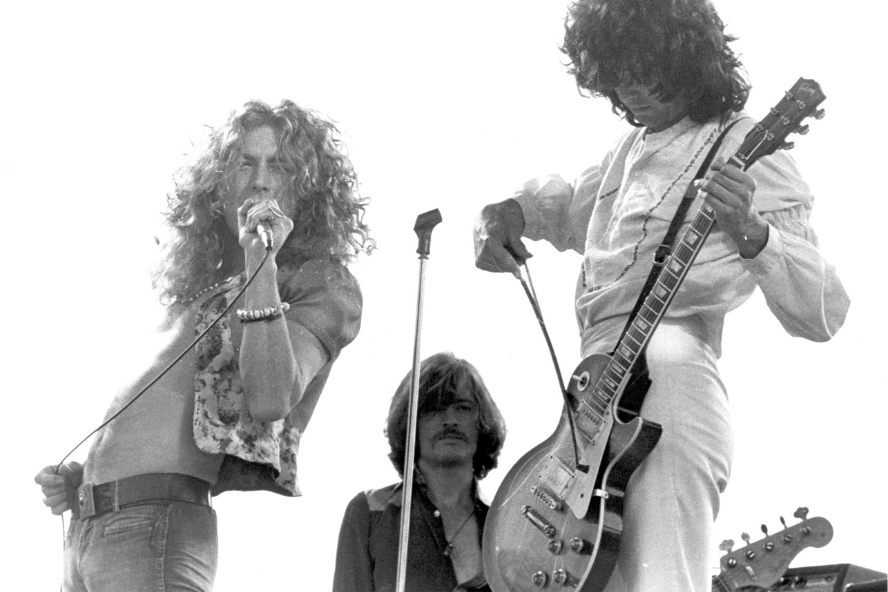 Robert Plant singing and Jimmy Page bowing his guitar in the foreground with John Paul Jones in the background during a '73 Led Zeppelin performance