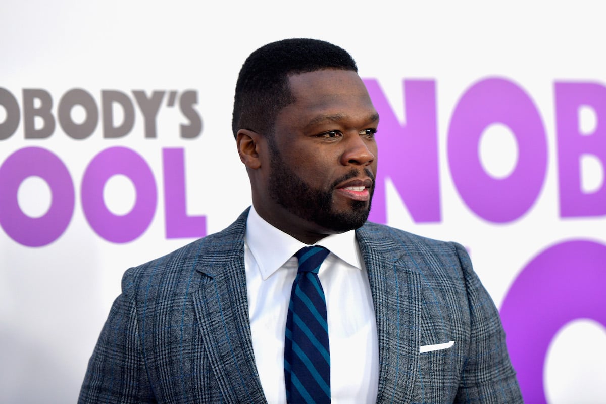 50 Cent stands on a red carpet wearing a grey suit.