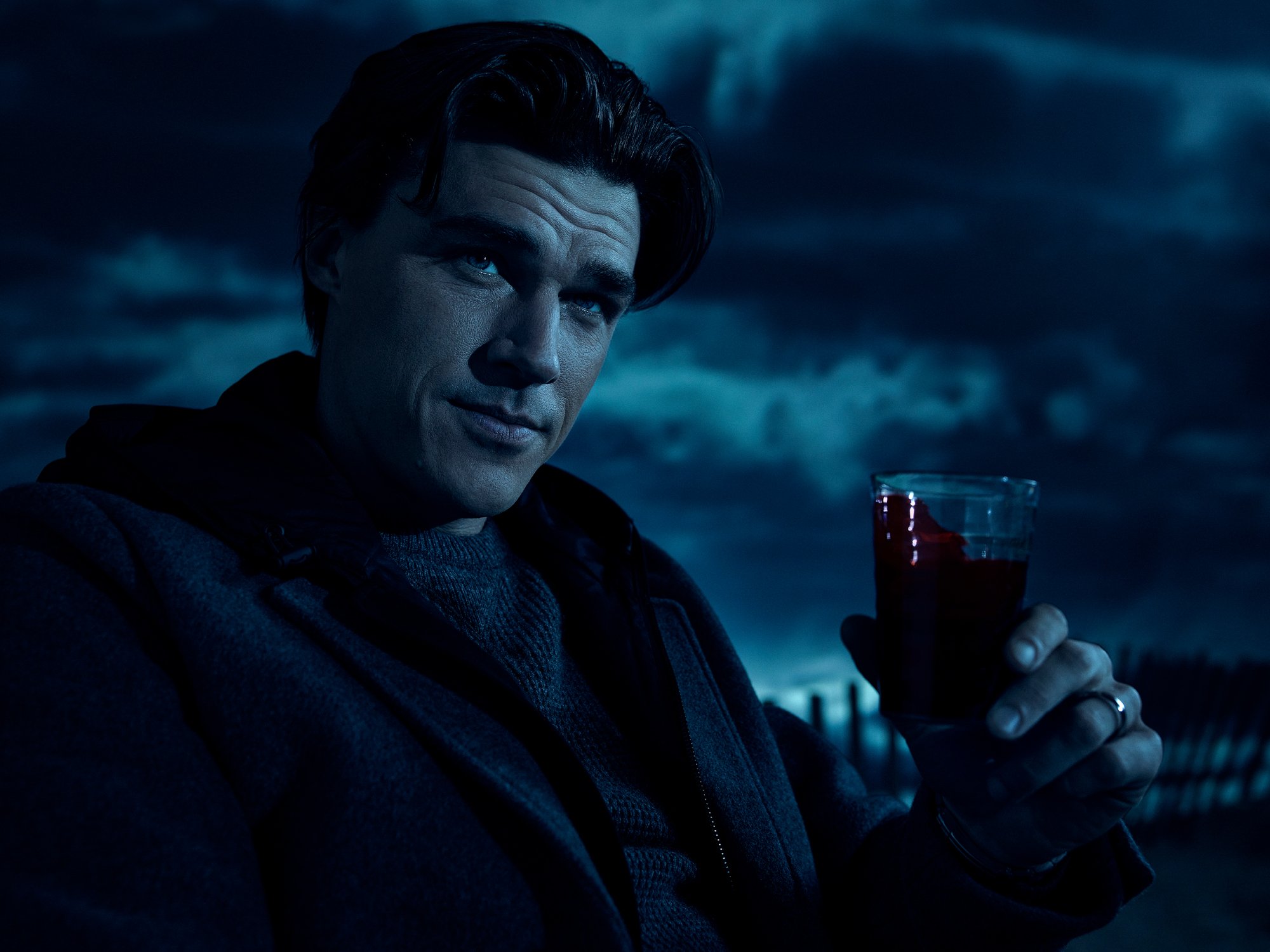 'American Horror Story' Season 10 image shows Finn Wittrock wearing a dark sweater and coat and holding a cup of blood. He looks like he's lounging and a cloudy sky is behind him.