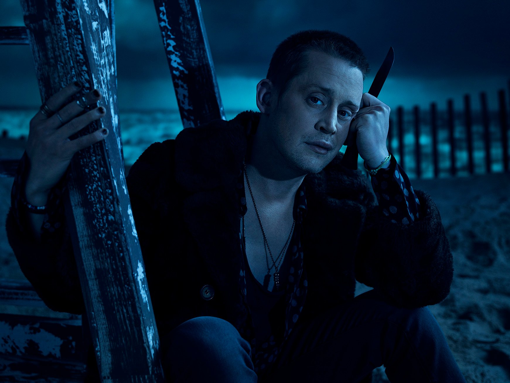 Macaulay Culkin as Mickey in 'American Horror Story' Season 10. He's sitting on the beach and holding onto a wooden pole while holding a knife in the other hand.
