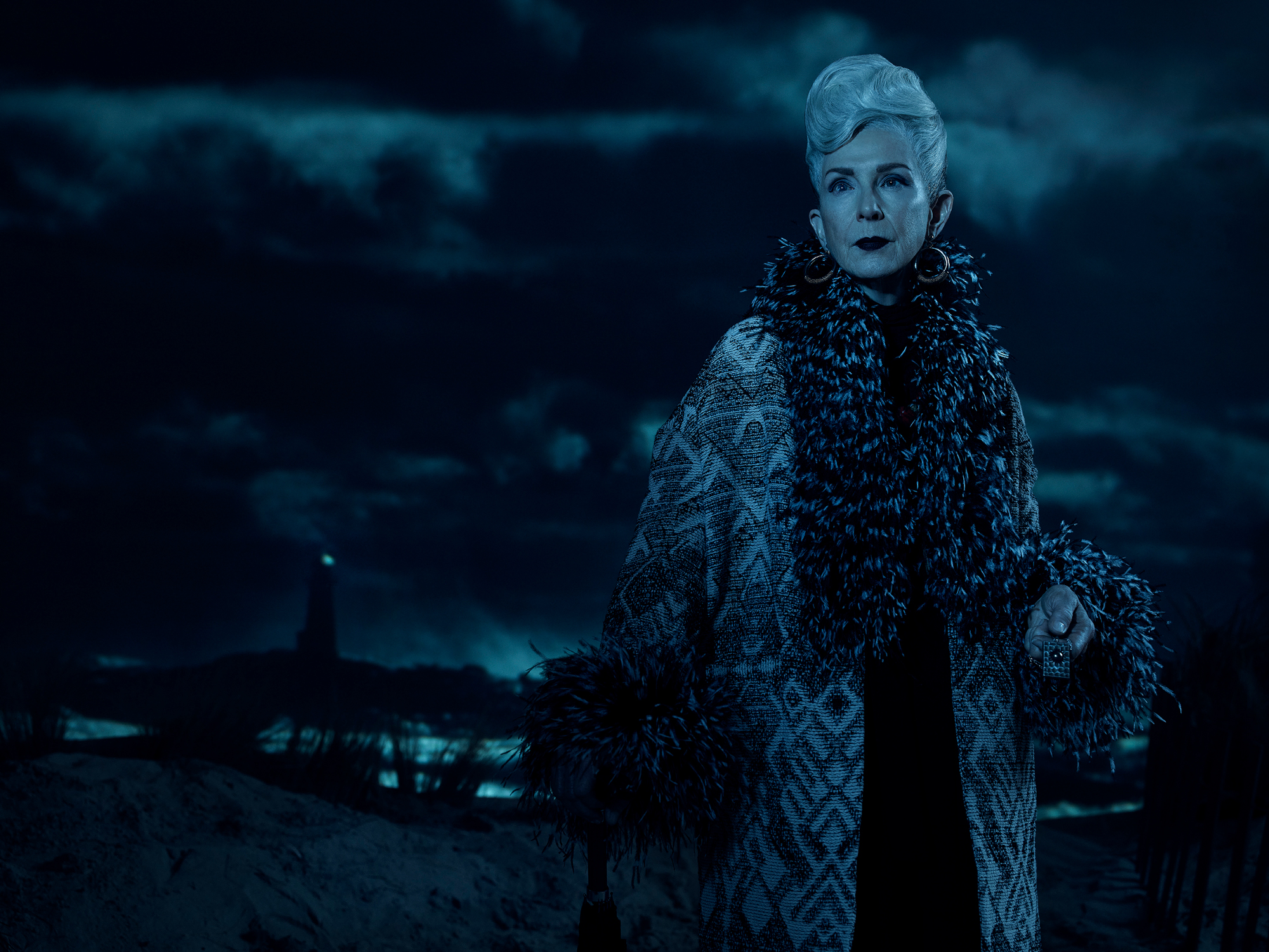 Frances Conroy as Belle Noir in 'American Horror Story' Season 10 Part 1. She's standing in front of a dark, cloudy sky with a lighthouse in the distance. She's wearing a big fur coat.