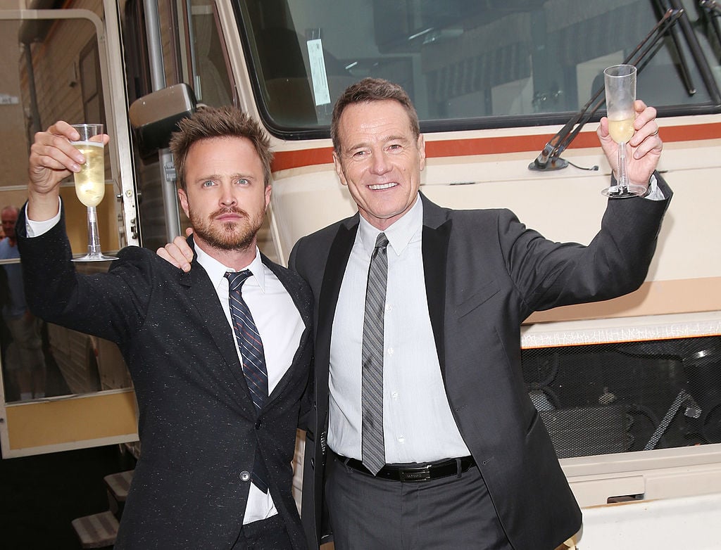 Aaron Paul and Bryan Cranston hold champagne toast glasses as they stand in front of the 'Breaking Bad' RV during a press event. Each are wearing a dark suit and tie with white shirts underneath and big smiles all-around.