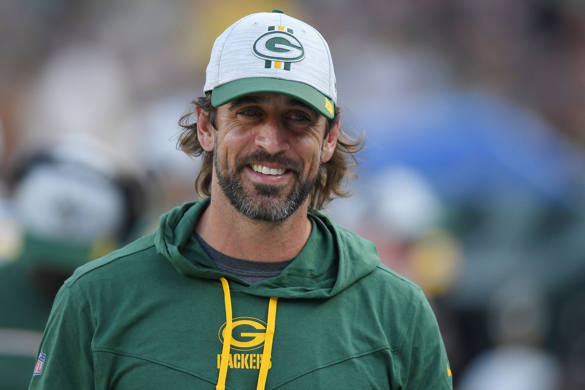 Aaron Rogers standing in front of blurred background in a green Packers hoodie and hat.