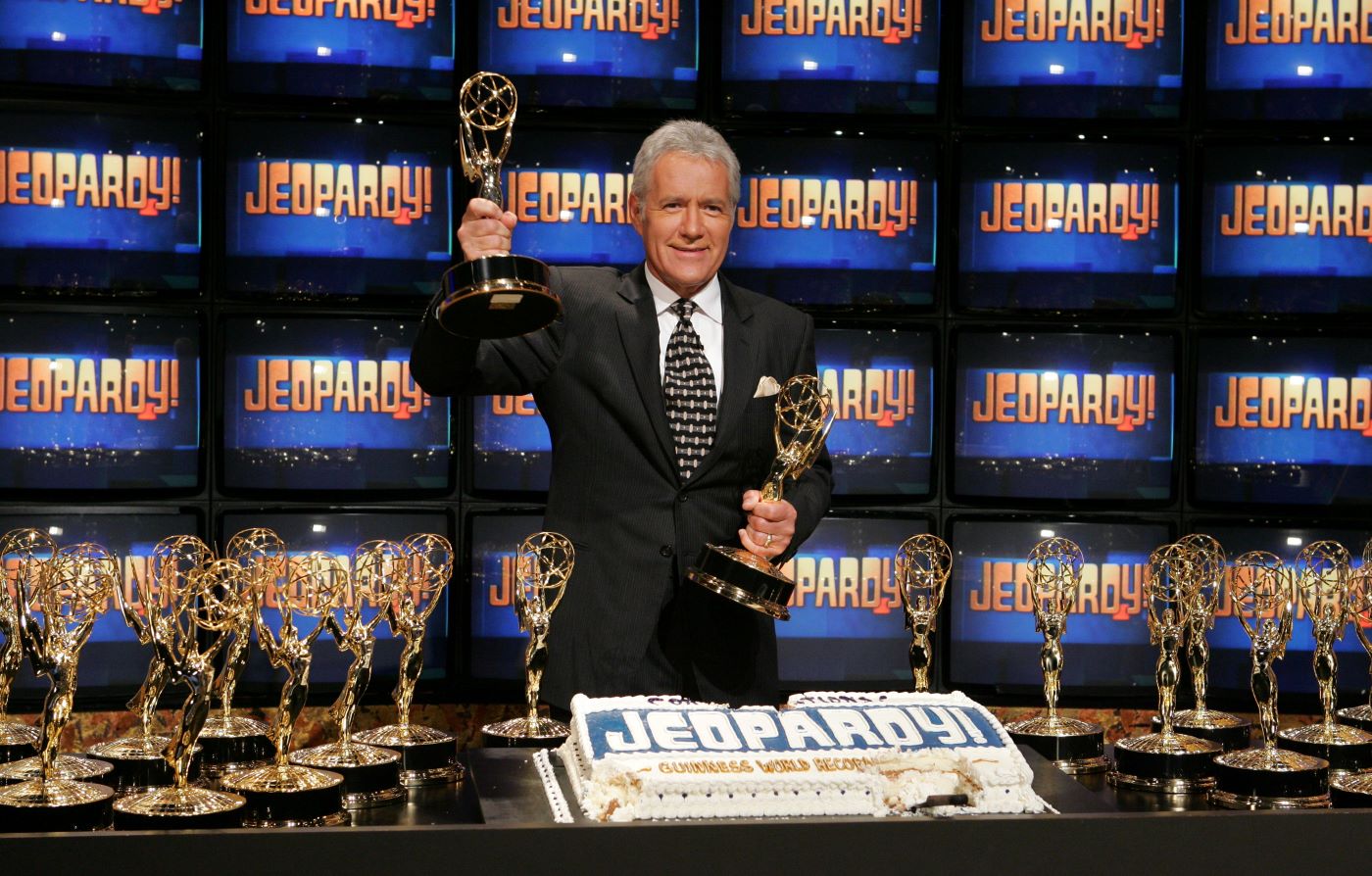 Alex Trebek from 'Jeopardy!' with the classic screens behind him surrounded by awards dressed in a suit and tie.