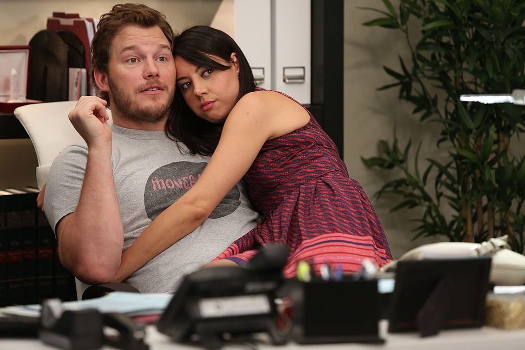 Chris Pratt as Andy, Aubrey Plaza as April Ludgate sit on the couch together. April has her arms around Andy.