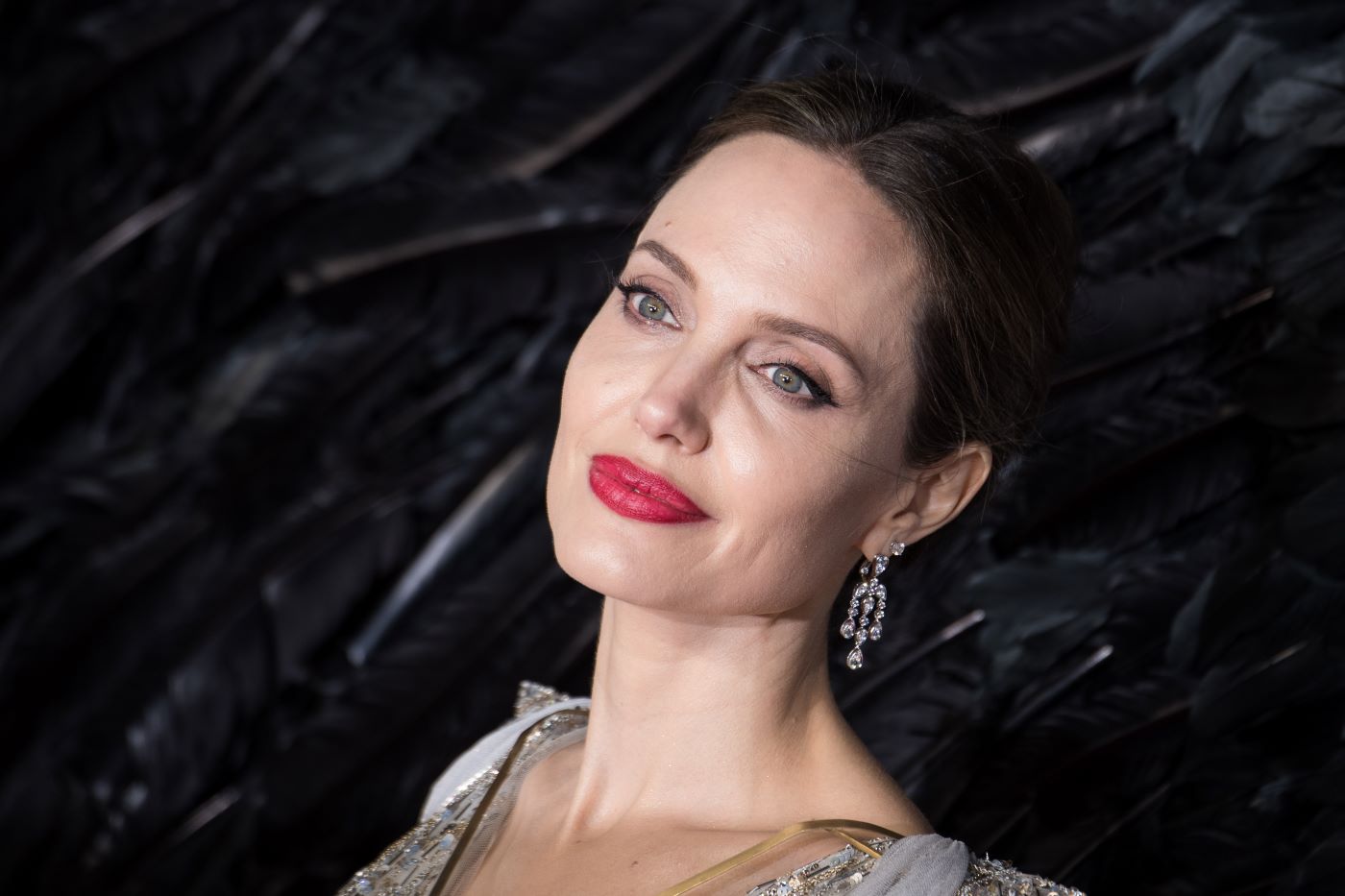 Angelina Jolie in a silver and gold top in front of a background of black feathers.