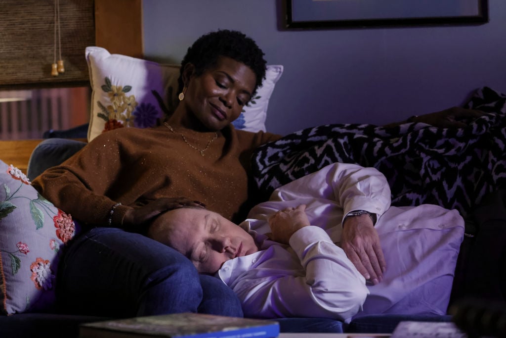 LaChanze as Anne looks down on James Spader as Raymond 'Red' Reddington as he lies his head in her lap, fast asleep. She's smiling.