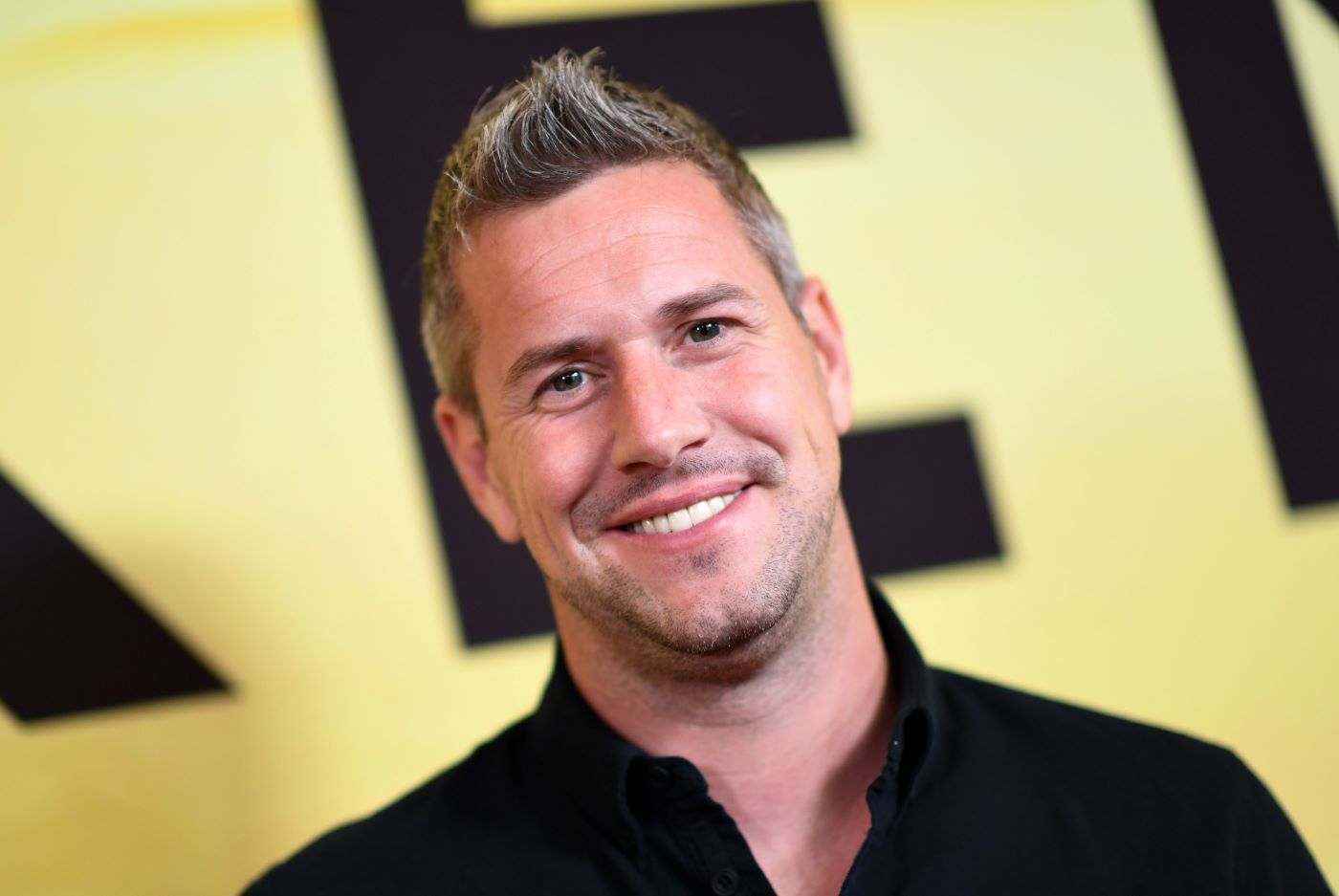 Ant Anstead wearing a black collared shirt in front of a yellow background with a black letter.