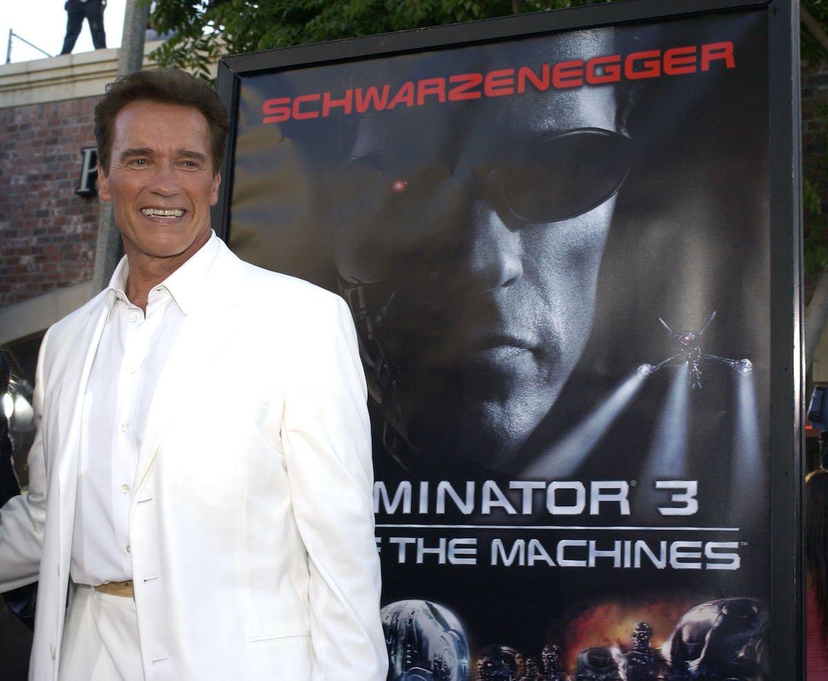 Arnold Schwarzenegger wears a white suit and poses beside artwork for ‘Terminator 3: Rise of the Machines’