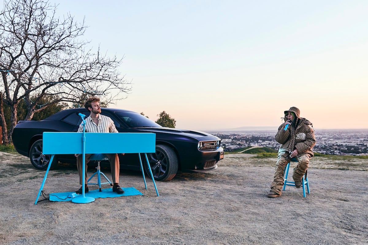 Finneas O'Connell and Billie Eilish perform outside next to a black car. Finneas plays a bright blue keyboard while Billie sits in a matching blue chair.