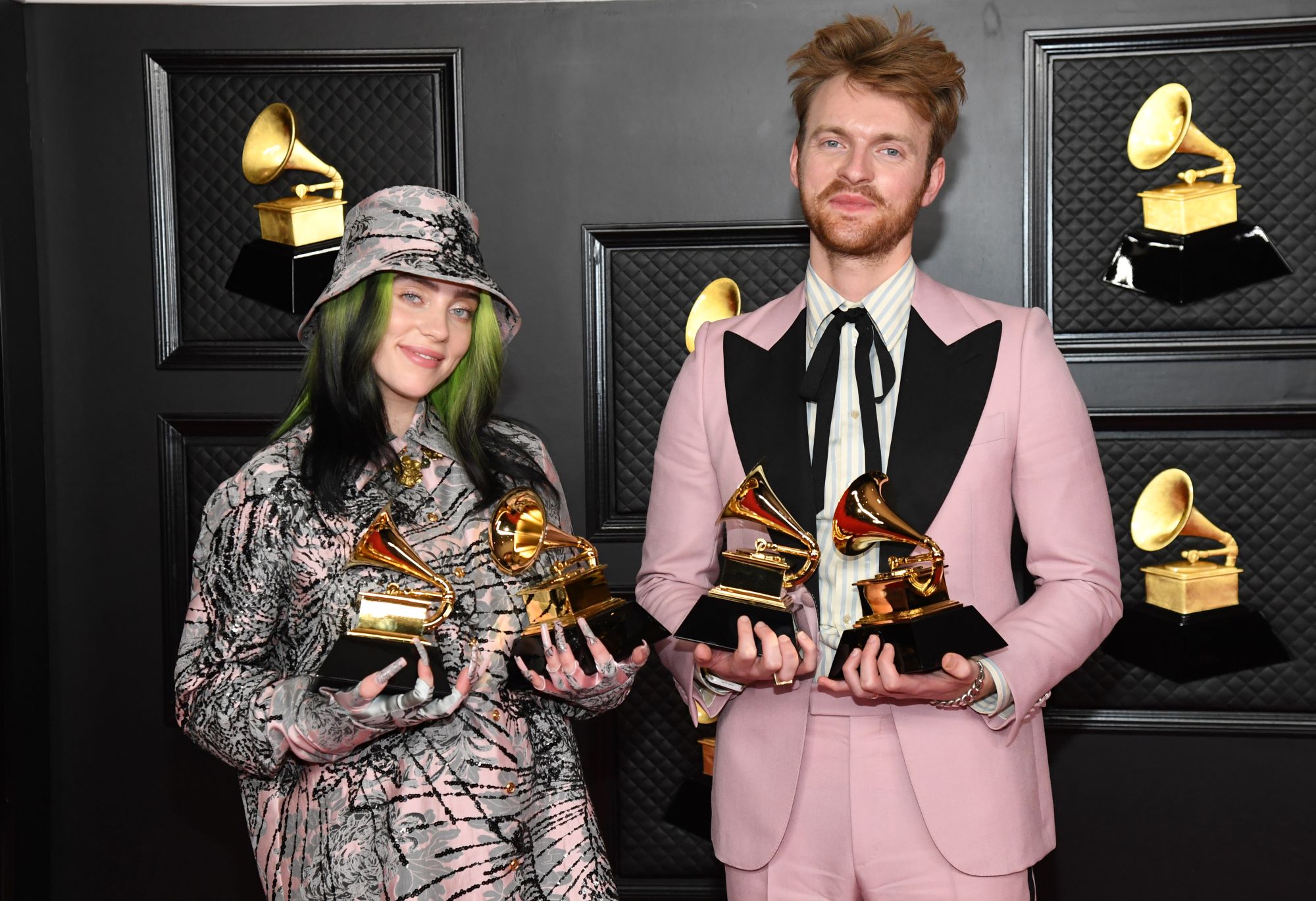 Billie Eilish dressed in a black grey and pink pattern outfit holding grammy awards next to her brother who is wearing a pink suit with a black collar and Texan bow tie with a white undershirt holding more grammy awards standing in front of a grey background.