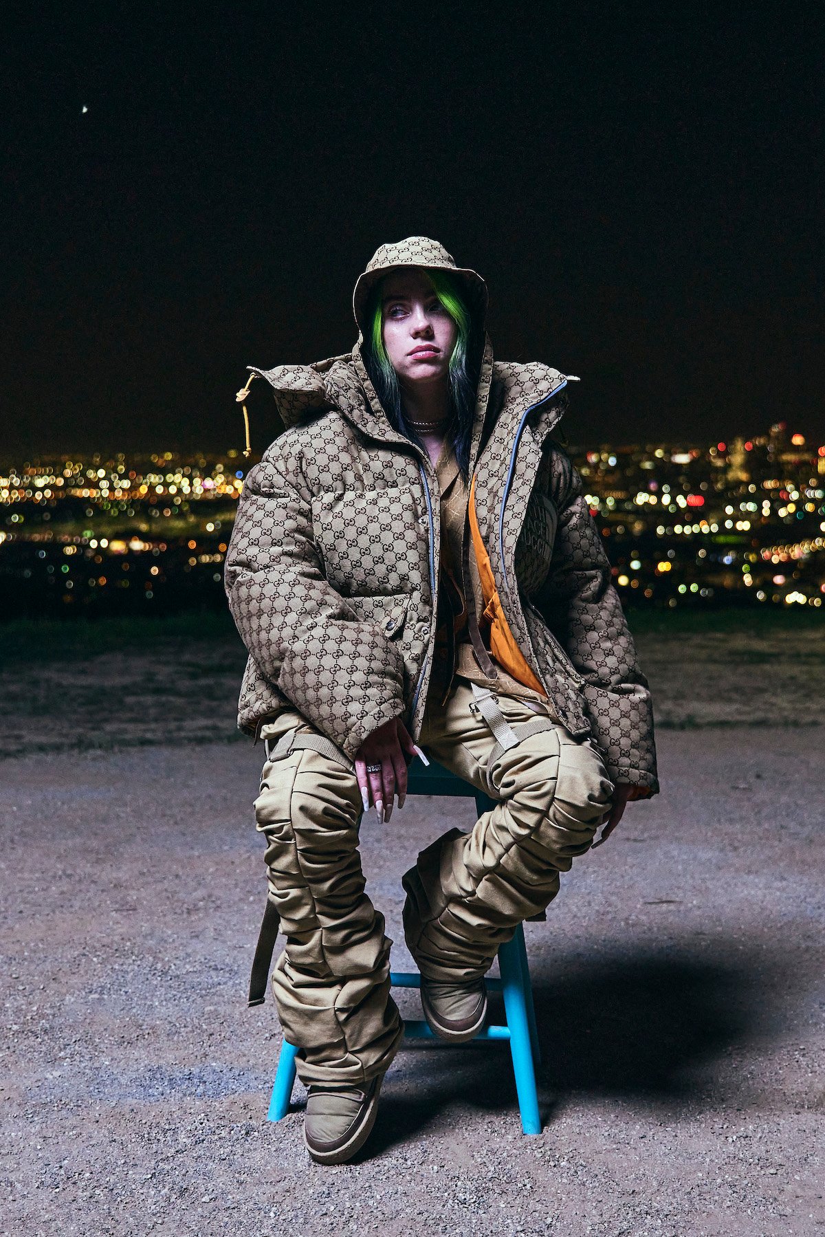 Billie Eilish alone in a chair facing the camera unsmiling, wearing an oversized puffy coat and baggy pants.