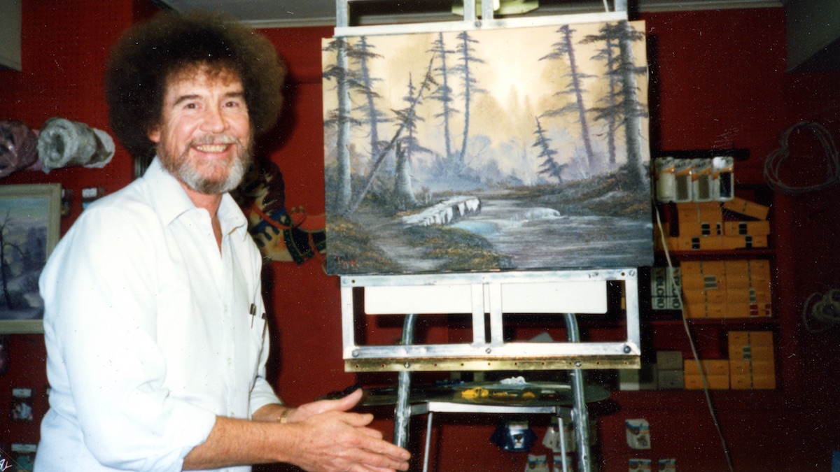 Bob Ross smiling at the camera next to a painting