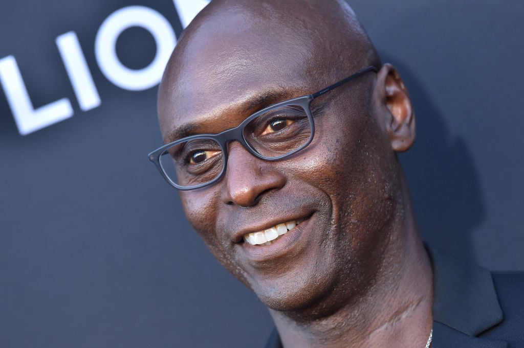 Lance Reddick smiles on the red carpet. He's wearing all black and glasses.