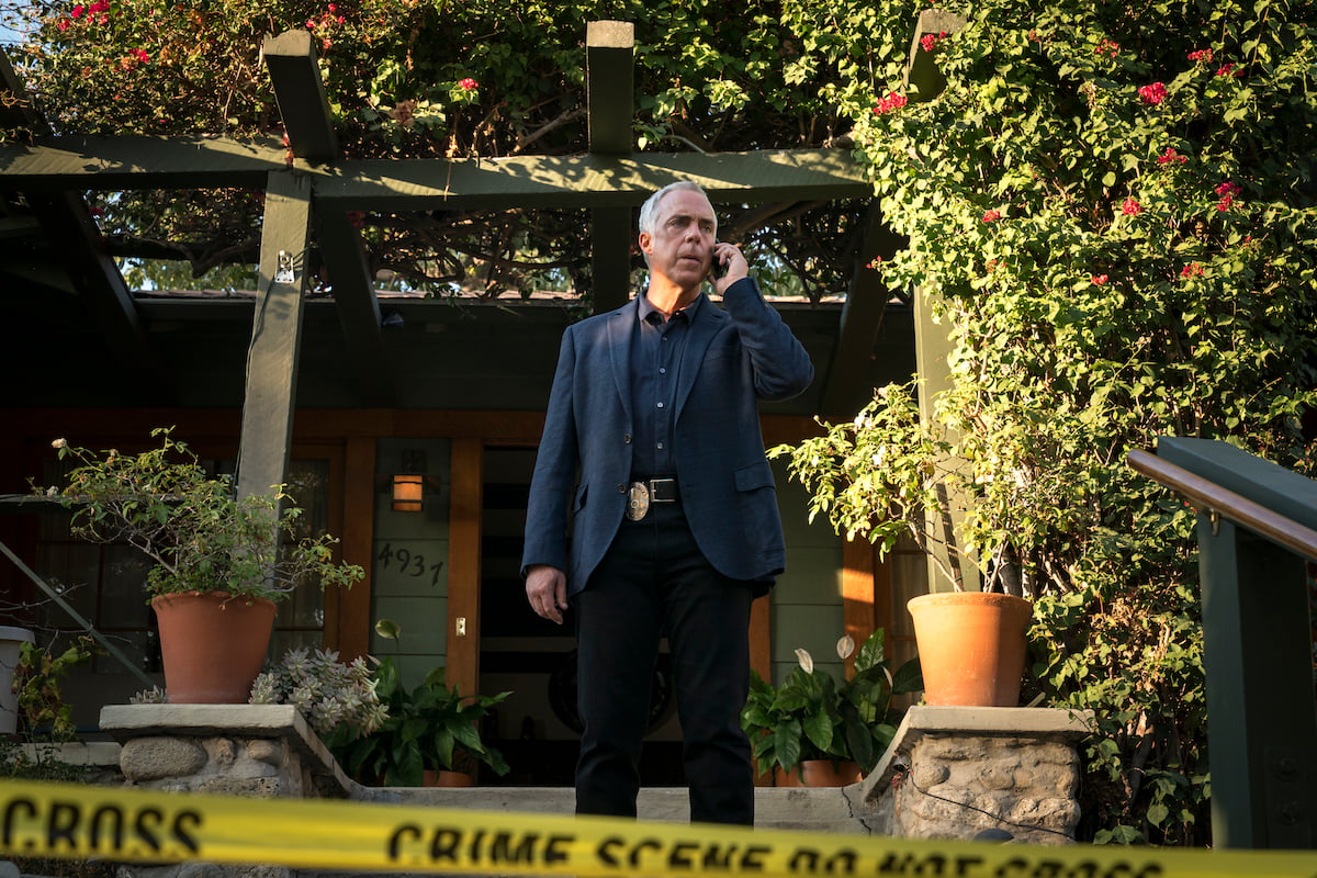 Titus Welliver as Detective Harry Bosch takes a call as he stands outside a crime scene. He's dressed in a navy suit and is surrounded by shrubs.