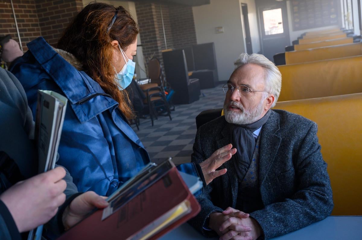 Director Liz Garbus and Commander Lawrence actor Bradley Whitford on set of 'The Handmaid's Tale' Season 4 Episode 10, 'The Wilderness.' Whitford sits in a wool coat, paisley suit vest, neck scarf, and glasses in a yellow booth at a diner in daytime. Whitford is playing Commander Joseph Lawrence in this scene and is being directed by Garbus. The floor is black and white and checkered and the walls are brick.
