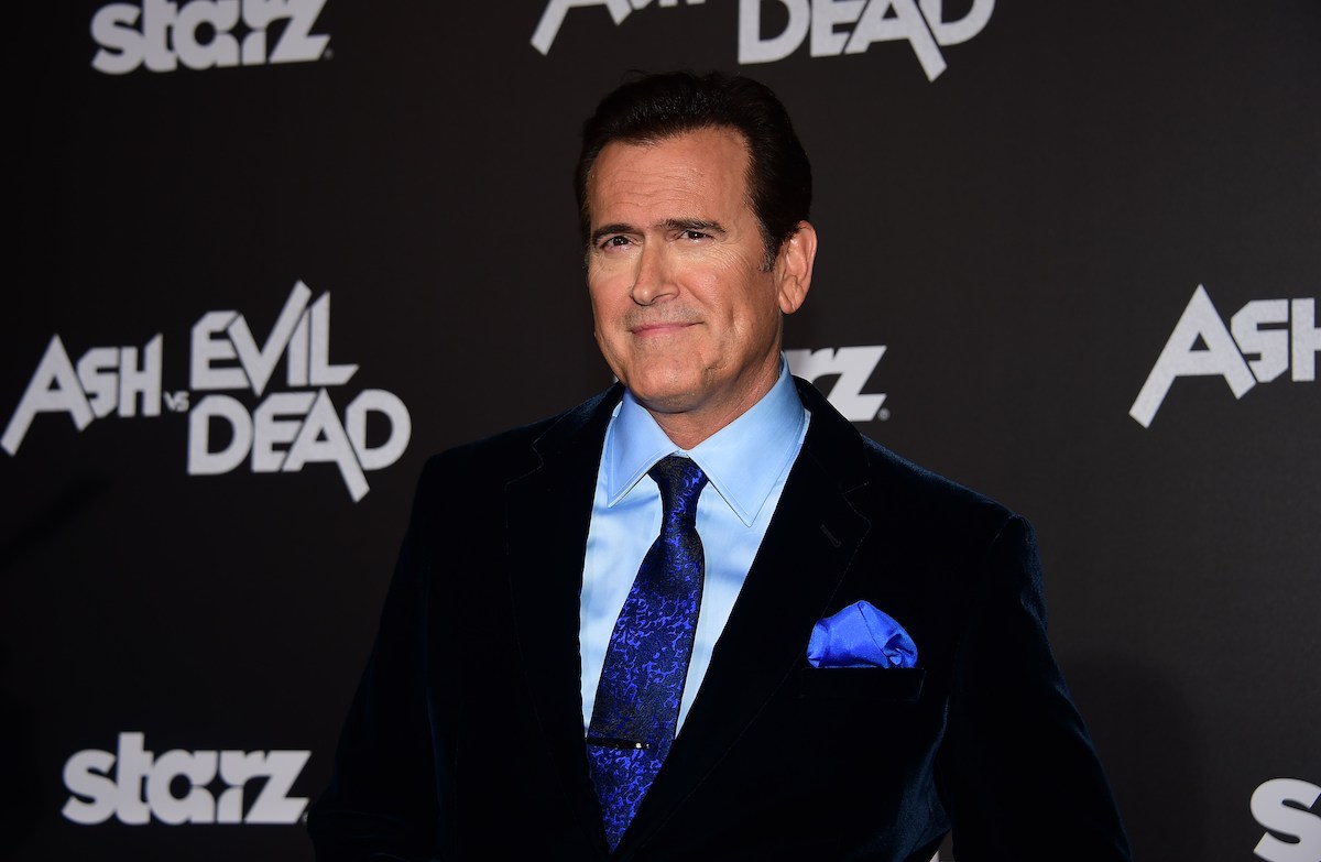 Bruce Campbell wears a suit and poses in front of the ‘Ash vs. Evil Dead’ logo