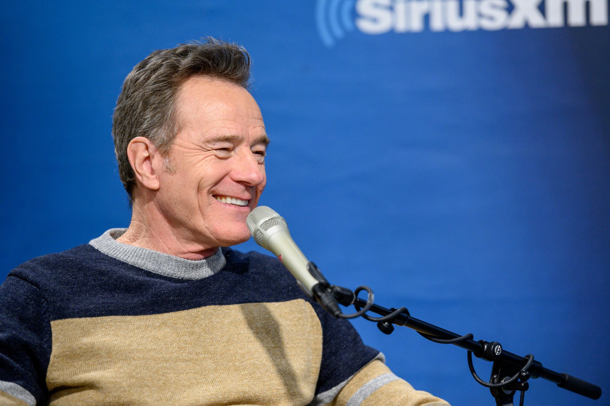 Breaking Bad star Bryan Cranston wearing a grey, navy, and biege sweater and smiling. There is a microphone in front of him and a SiriusXM Studios wall behind him.