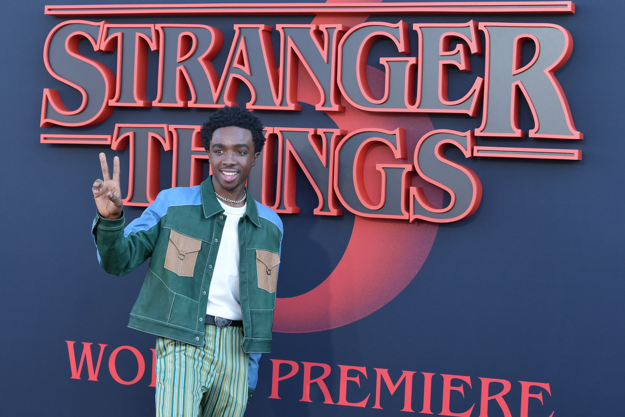 Stranger Things Season 4 star Caleb McLaughlin holds up a peace sign in front of a wall with the show's title on it. He's wearing a green jacket with brown pockets on the front and blue shoulders.
