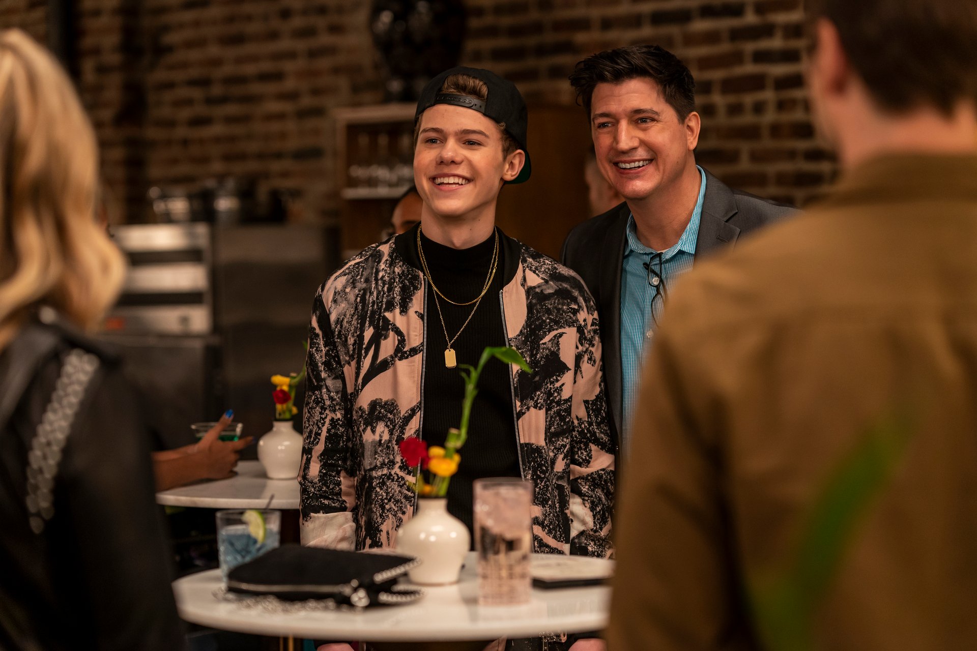 Case Walker and Ken Marino stand in front of a table with a vase of flowers and a drink.