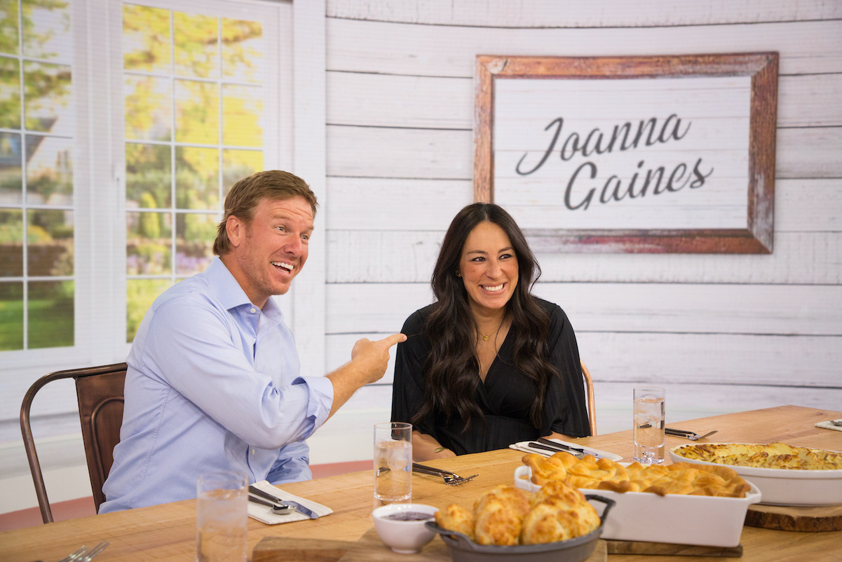 Chip smiles and points to Joanna Gaines while the two sit at a table
