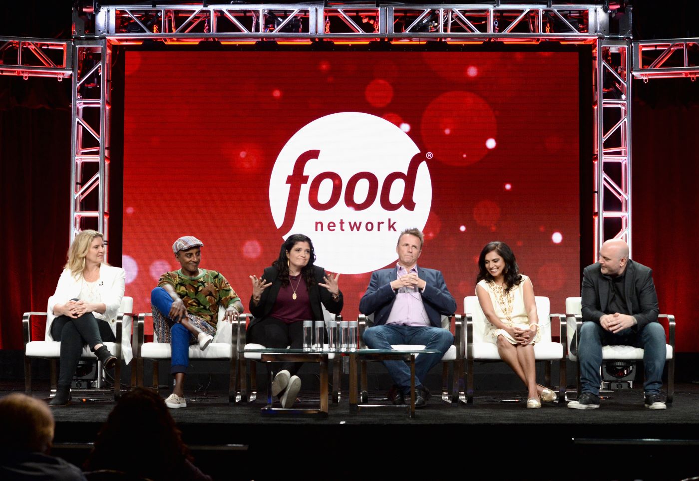 6 'Chopped' judges sitting on a stage dressed professionally in front of an industrially framed red screen with food network printed in white on it.