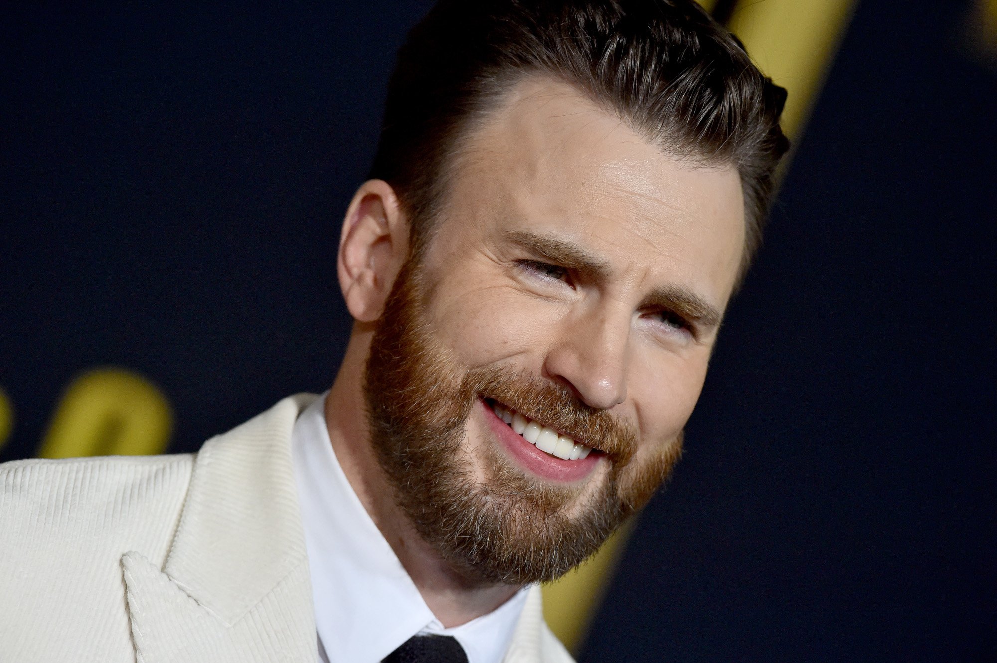 Chris Evans, who does not voice Steve Rogers in Marvel's What If...? TV series,. He's wearing a white suit and has his beard grown out. He's smiling.
