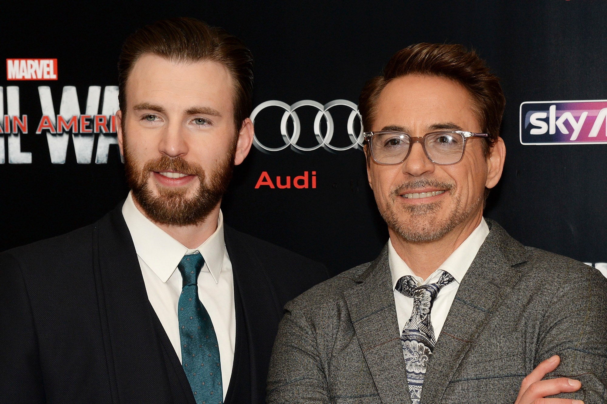 Chris Evans stands next to Robert Downey Jr. at the premiere for 'Captain America: Civil War.' Evans wears a black suit, white shirt, and blue tie. Downey wears a grey suit and tie with a white shirt.