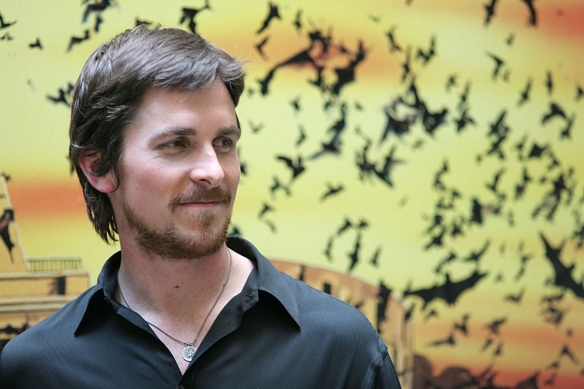 Christian Bale smiles in a dark shirt in front of ‘Batman Begins’ artwork